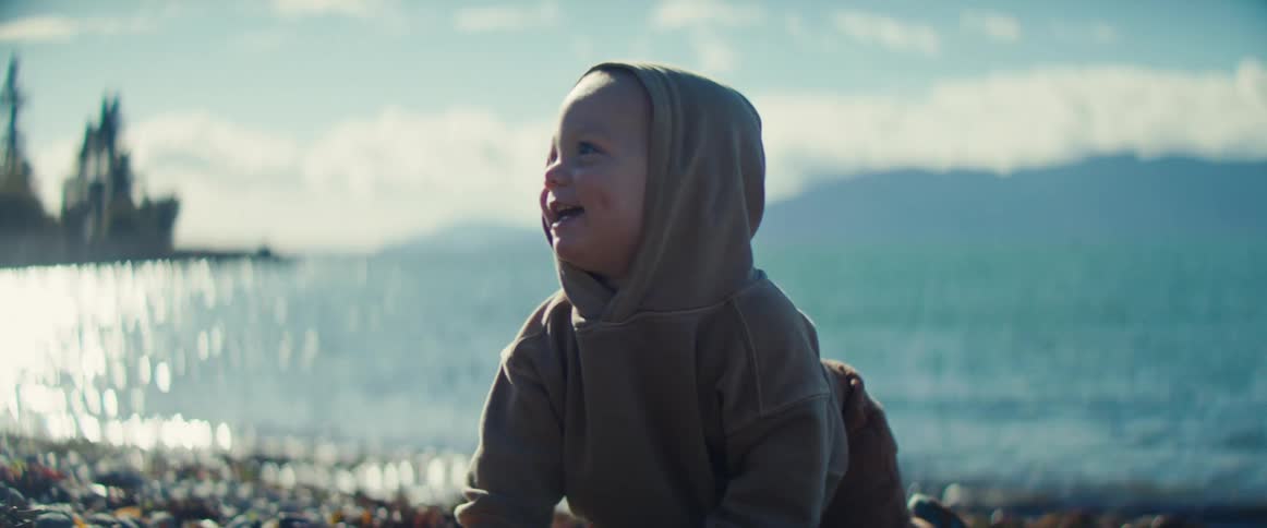A baby at the edge of the beach picking up a stone and putting it in their mouth