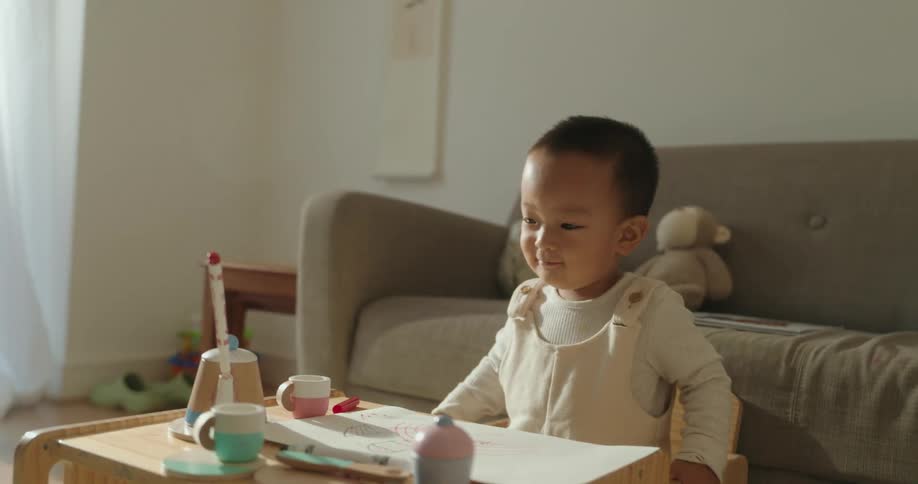 A happy toddler wearing light overalls plays with toys at a small wooden table in a bright sunny living room as a womans arm reaches in from the side to help him