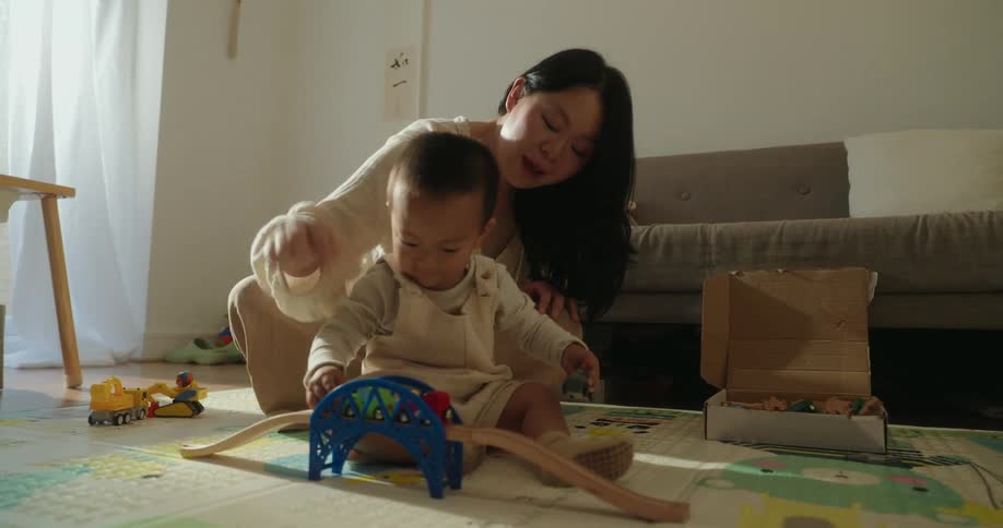 Asian mother supervising her baby playing with toys while sitting on the floor in the living room