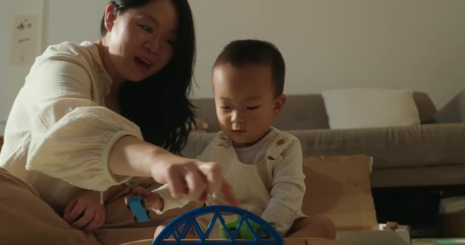 A mother and her young child are seated on the floor together playing with a wooden toy train setthe mother leans in to interact with and guide the child who is focused on pushing a small toy car across a blue toy bridge