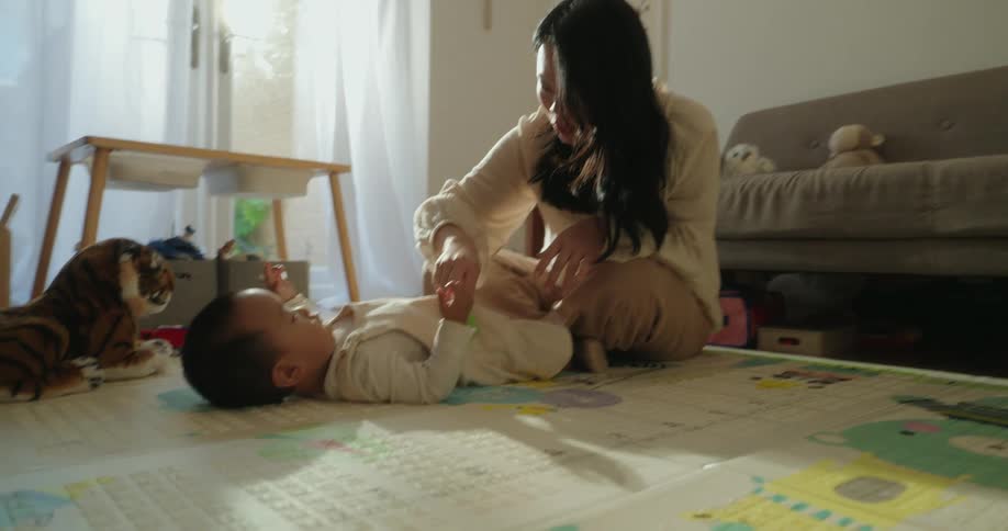 A woman plays on a colorful floor mat while a baby lies on their back reaching toward a toy