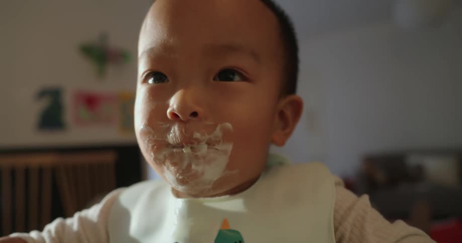 The face of a baby with white food smeared on his mouth as he eats and moves his head towards the food
