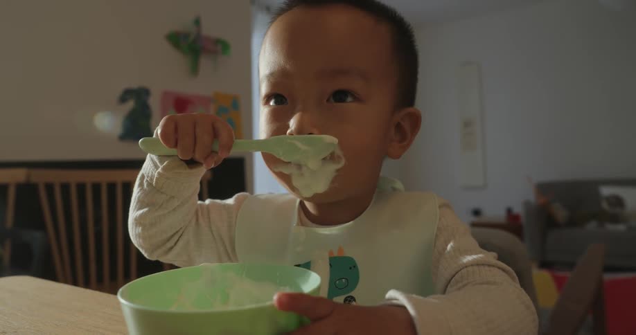 A toddler eats with a spoon from a bowl at a table in a softly lit home