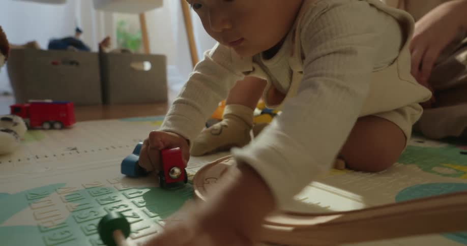 A mother and her baby are captured in a closeup sitting on the floor in a bright sunlit room with the toddler intently playing with colorful wooden toys and train tracks on an activity mat while the mother hovers nearby