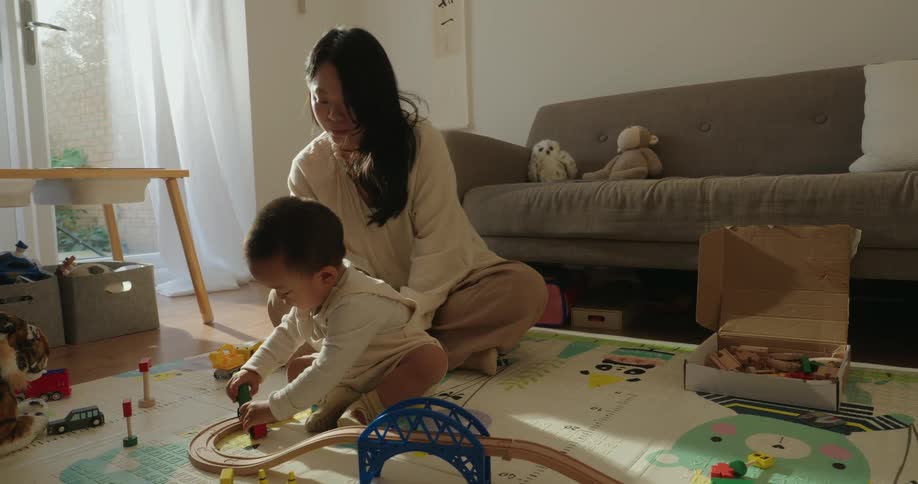 Mother sits on the living room floor with her son and watches him play with a toy vehicle