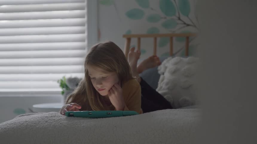 A young girl lies on a bed absorbed in a tablet near a bright window with blinds