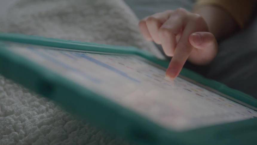 A small hand taps a tablet in a teal case on a quilted blanket