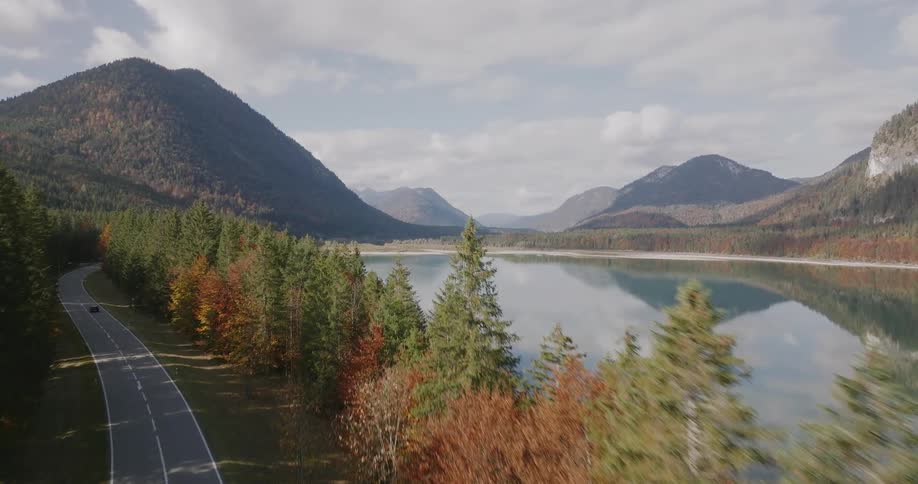 Car travels on a forested mountain road as small boats sail on the mountain valley lake