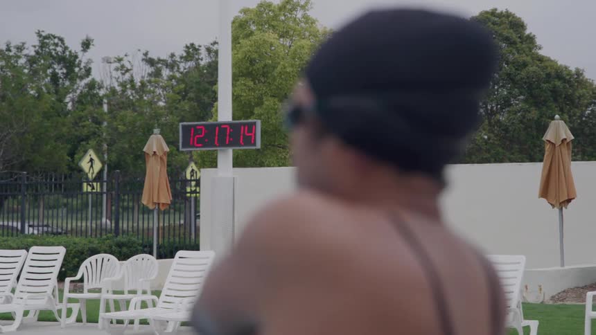 A blurry figure stretches while seated preparing to swim as a sharply focused digital clock in the background reads 121714