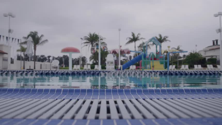 A quiet outdoor pool sits under an overcast sky with palm trees and a colorful splash playground in the background