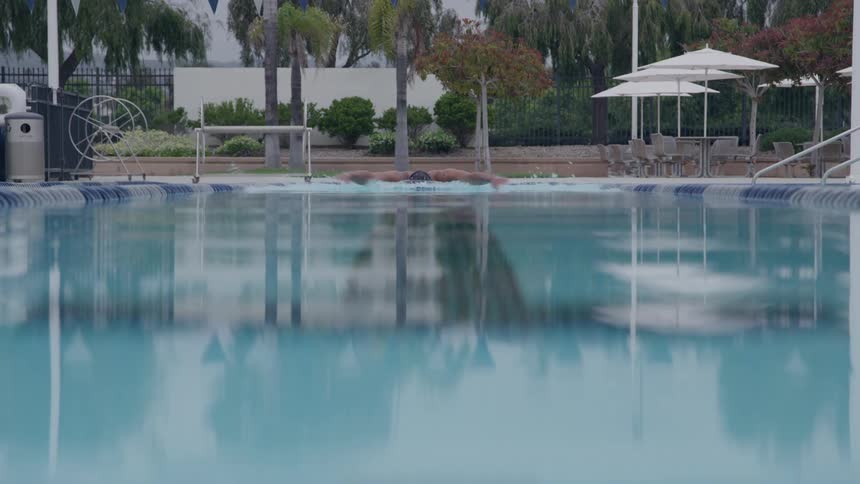 A swimmer wearing dark goggles propels themselves through the bright blue water of an outdoor pool using the butterfly stroke creating significant splash in the foreground while a resortlike area with white umbrellas and palm trees on the background