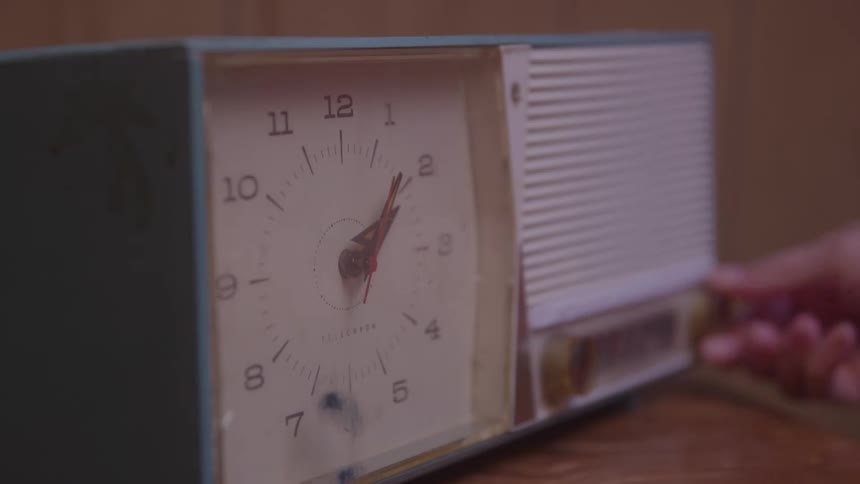 A retrostyle analog clock radio with a light blue casing sitting on a wooden surface as a persons hand enters the frame to delicately turn a knob or dial on the side of the device adjusting the radio setting
