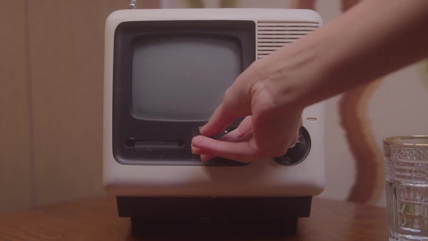 A small vintage offwhite television sits on a wooden surface against a retro patterned wall a hand reaches in to turn the volume dial but the television remains off