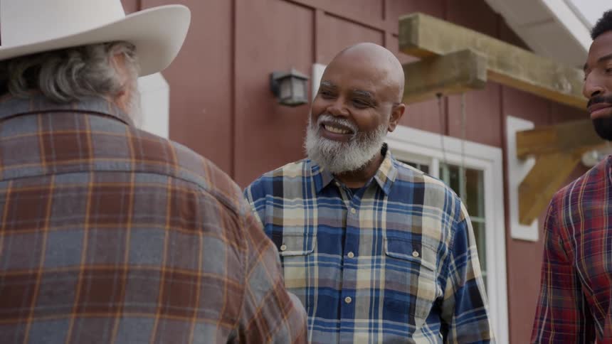 A smiling man in a blue plaid shirt chats outdoors with two others by a red building