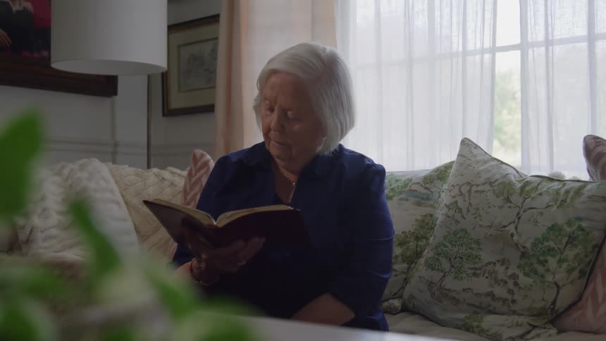 An elderly woman sitting on a couch reading a book