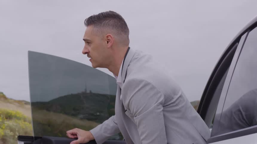 The video begins with a closeup on the side of a white car possibly a tesla model y parked on a road with the door opening to reveal a man in a light gray suit getting outthe man who has a closecropped haircut fully exits the vehicle and stands