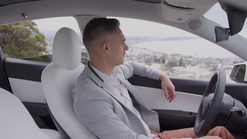 The video shows a man with a groomed haircut and grey jacket sitting in the drivers seat of a modern car with white interior looking out the window with a pensive expression the background visible through the car window is a scenic coastal or hillside