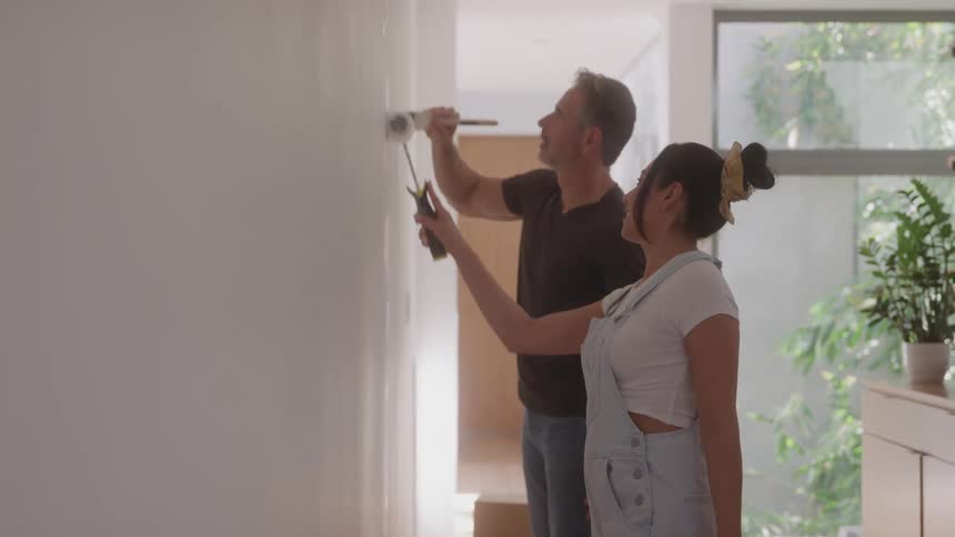 A man and a woman are painting a white wall together in a bright modern room with large windows the man uses a brush near the top while the woman uses a paint roller closer to the middle