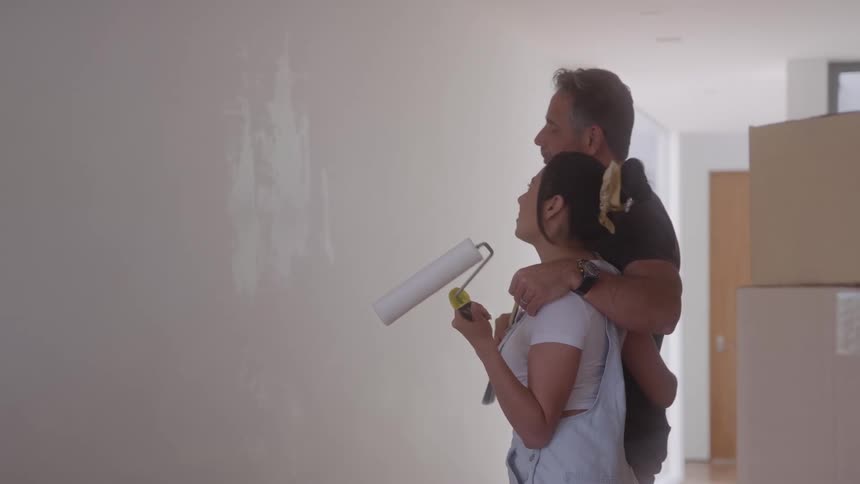 A couple talking and holding paintbrushes and a paint roller looking at a white wall