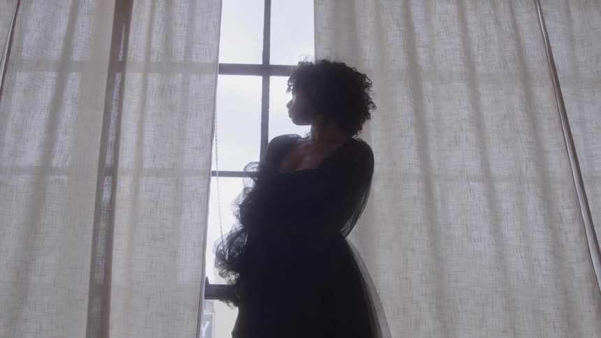 A young woman with curly hair is shown in silhouette standing in front of a window and looking to the side she is framed by sheer white curtains and the bright natural light pouring in from outside