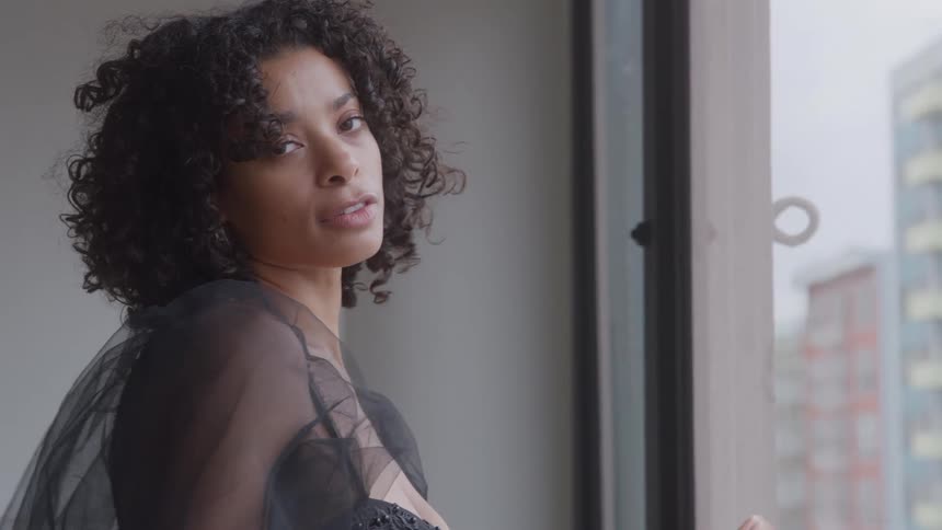 A woman with curly hair wearing a dark sheer top stands near a large window looking out at a city skyline she turns her head to look toward the camera with a focused expression