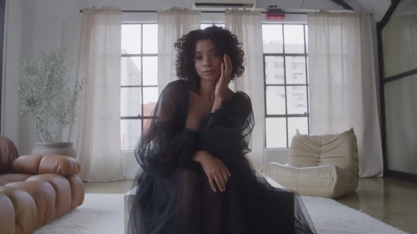 A woman with curly hair in a sheer black tulle gown sits gracefully on a chair in a sunlit loft with tall windows neutral lounge seating and a white rug