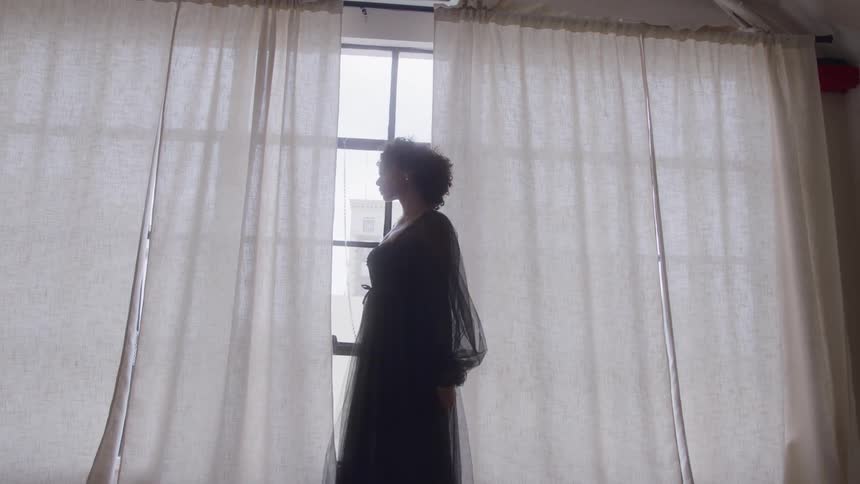 A young black woman wearing a dress stands beside the window curtain at midday