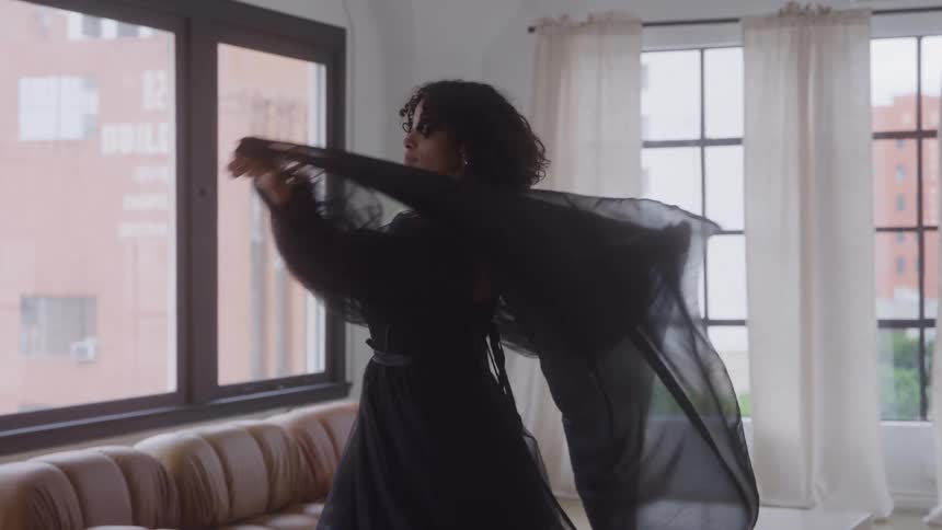 A young woman wearing a black dress spins around in the living room at midday