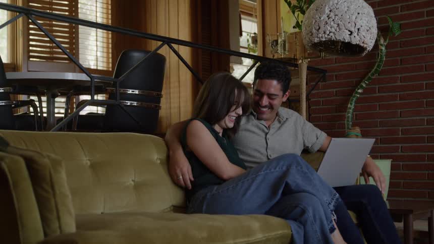 A couple sits together laughing while looking at a laptop on a cozy living room