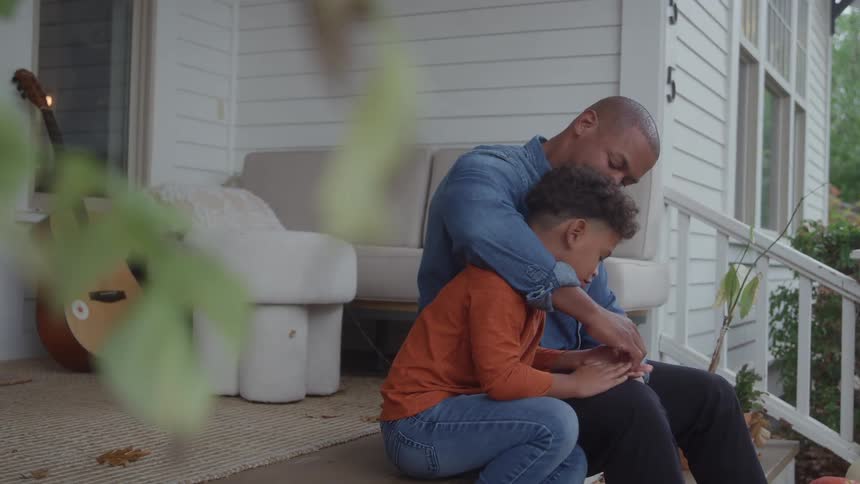 A father hugs son warmly while sitting together on the porch outside their home