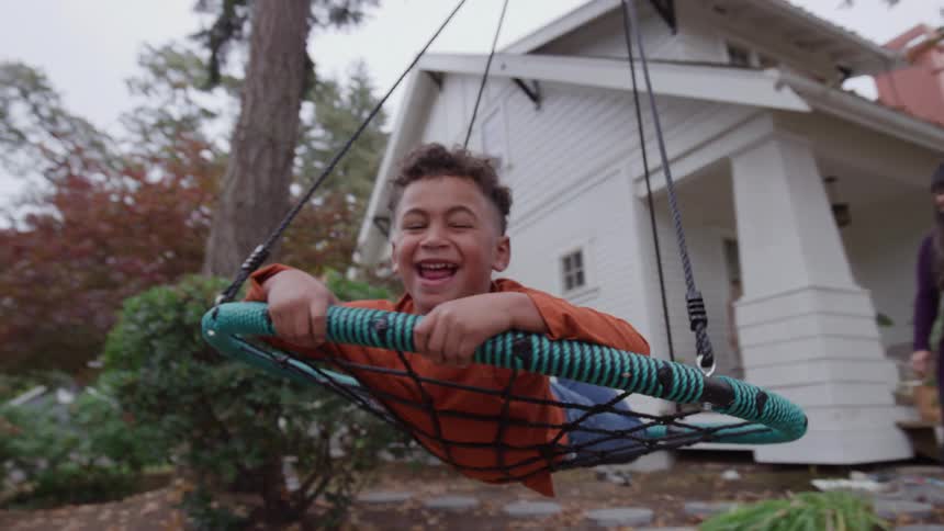 A happy boy lies on a large round net swing while his parents push him from behind in the yard of a white house he is smiling and enjoying the ride as the swing moves high through the air