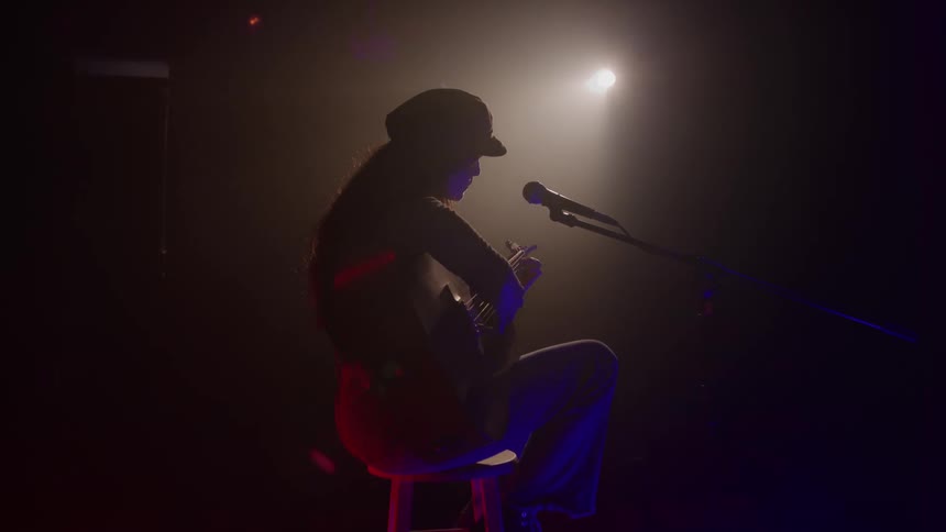 A female musician sings and plays guitar under a spotlight on a dark stage