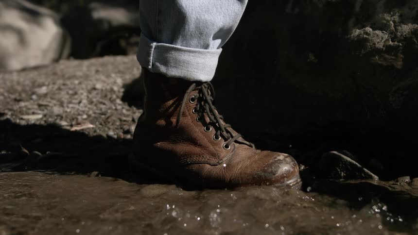 A worn leather boot steps into a shallow puddle on rocky ground