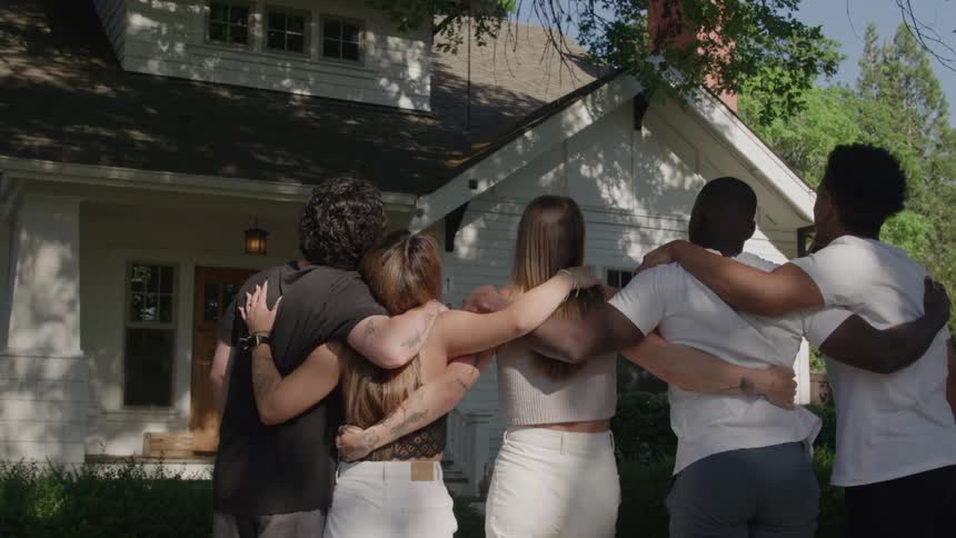 Group of friends with arms around each other look at a house lit by daylight