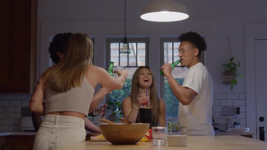 A group of friends in a cozy kitchen clink drinks around a wooden island under a warm pendant light