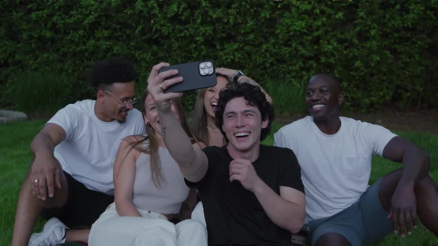A group of friends sitting on a backyard lawn smile and laugh as they pose for a selfie together