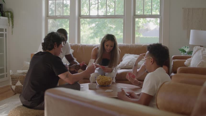 A group of friends are playing a card game at a table in a living room and they are smiling