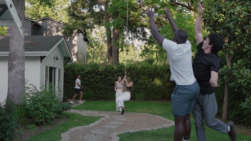 Three young men play baseball in the backyard with one tossing the ball into the air and another catching it while two young women enjoy a rope swing one being pushed from behind by the other