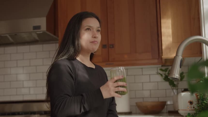 Woman drinks a green juice in a kitchen lit by natural light