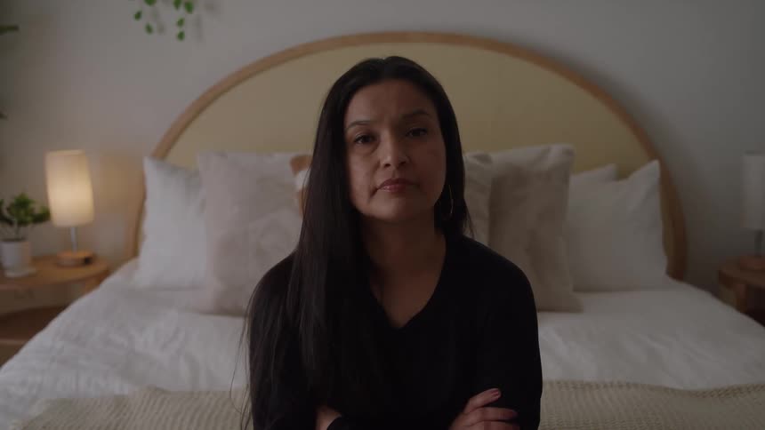 A woman with a sad look sits on the bottom edge of a bed her arms crossed then she lifts her head and looks forward