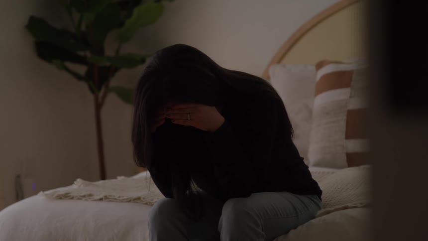 Woman sits on the bed covering her face in a moment of sadness