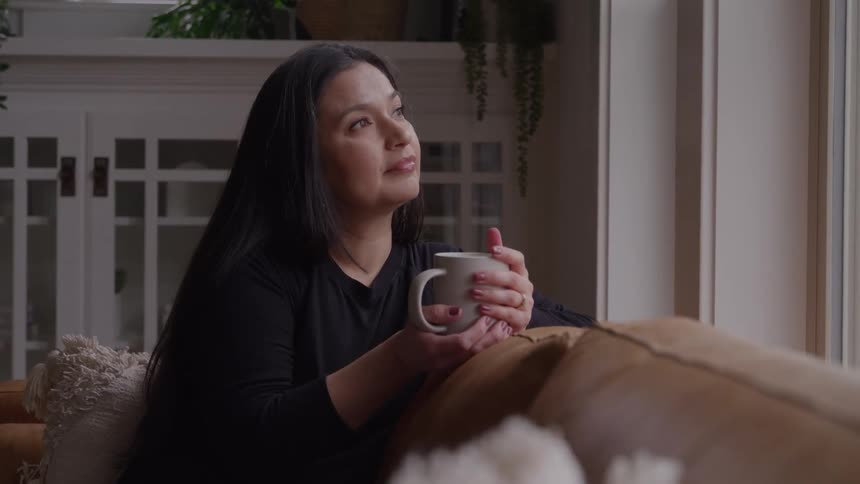 A woman with a sad look sitting on a couch turned to its back as she leans on the back of the couch her hands around a mug then lets out a wistful sigh as she looks toward the window