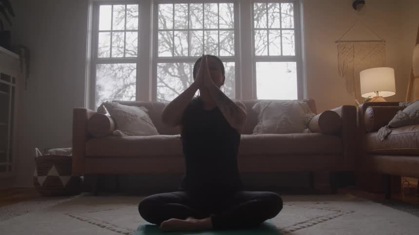 A woman wearing a black tank top is sitting on the floor of a living room doing yoga raising her arms upwards