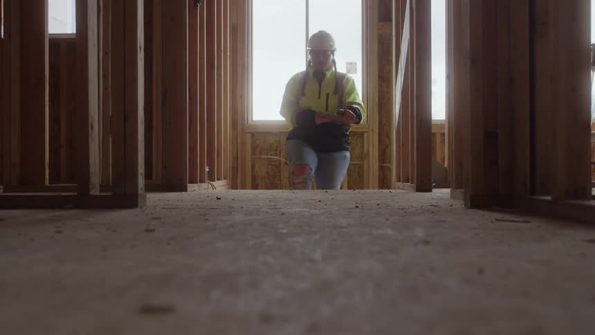 A woman wearing a safety vest goggles and a hard hat walks into a room under construction looking around