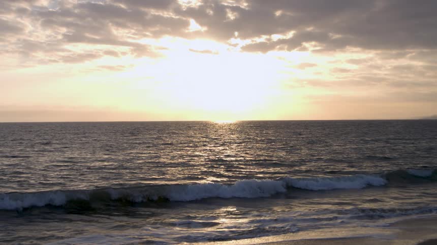 A single wave rolls toward the shore in slow motion as the bright sun sets on the horizon casting a shimmering reflection across the water under a cloudy sky
