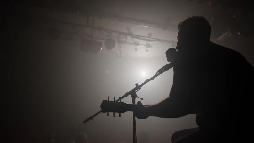 A musician is captured in a strong silhouette on a dimly lit stage hunched over an acoustic guitar and leaning into a microphone with atmospheric stage fog catching the light behind the performer