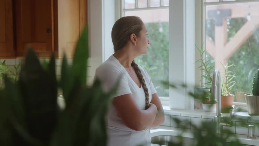 A woman with her arms crossed stands quietly at a bright kitchen window gazing outward with a pensive expression as she is softly lit by natural light with various houseplants visible in the immediate foreground and on the windowsill