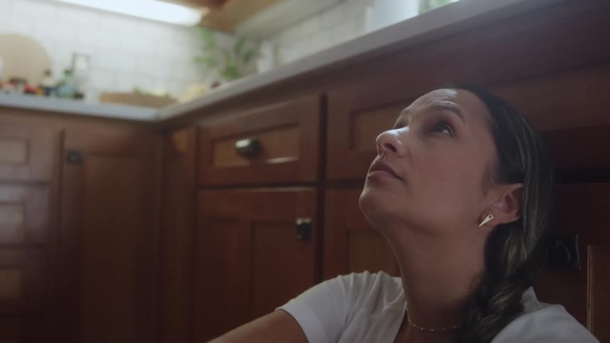 A lowangle shot shows a woman with a braided ponytail looking up while sitting in a kitchen