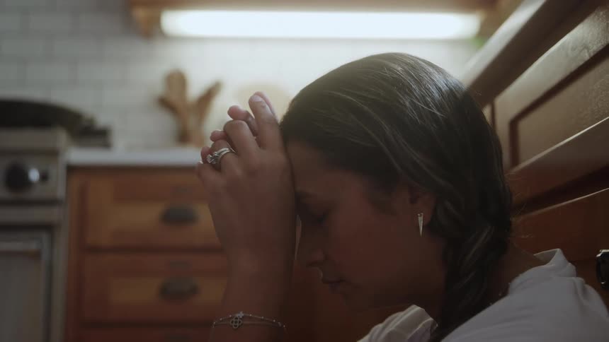 Woman sitting on the kitchen floor looks up and places her hands on her forehead praying