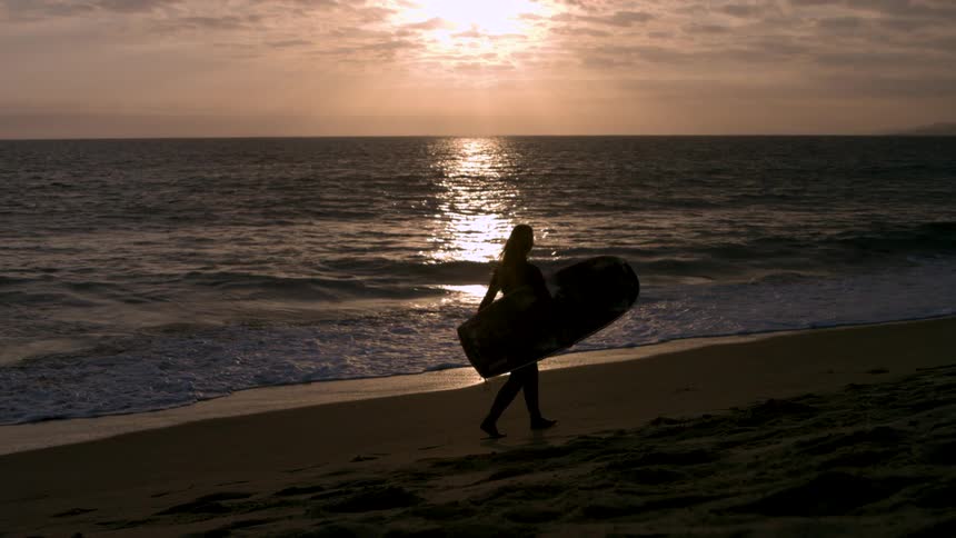 A woman carries a surfboard on the beach at sunset